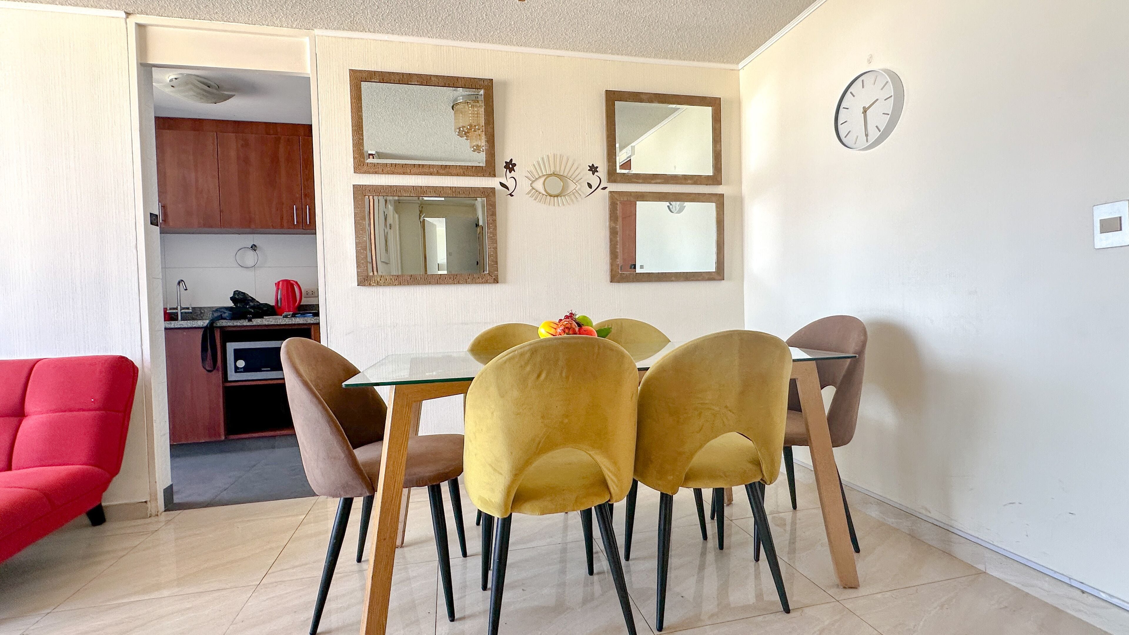Un comedor con una mesa y cuatro sillas, un sillón rojo y una cocina al fondo.