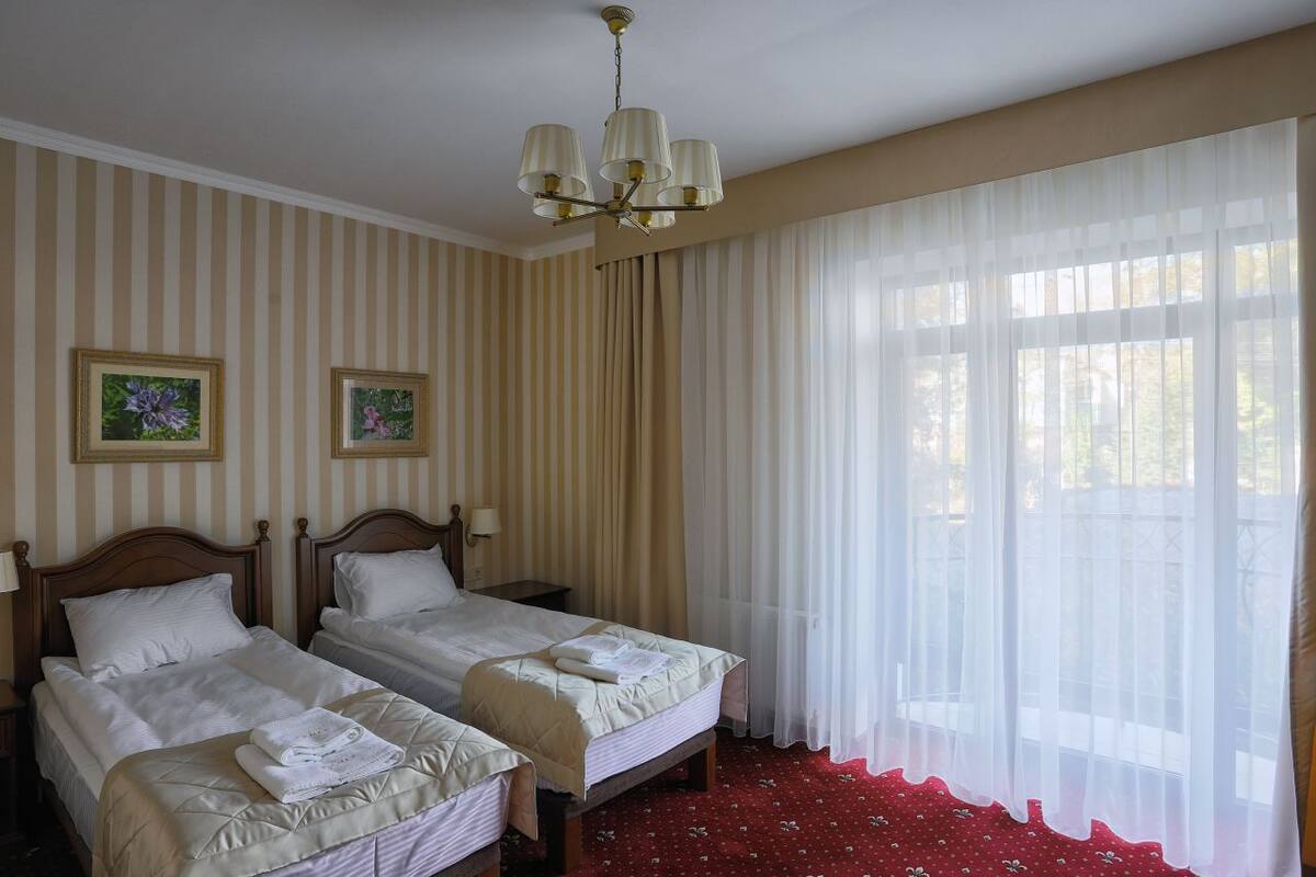 A hotel room with two single beds, a chandelier, striped wallpaper, and a large window with sheer curtains.
