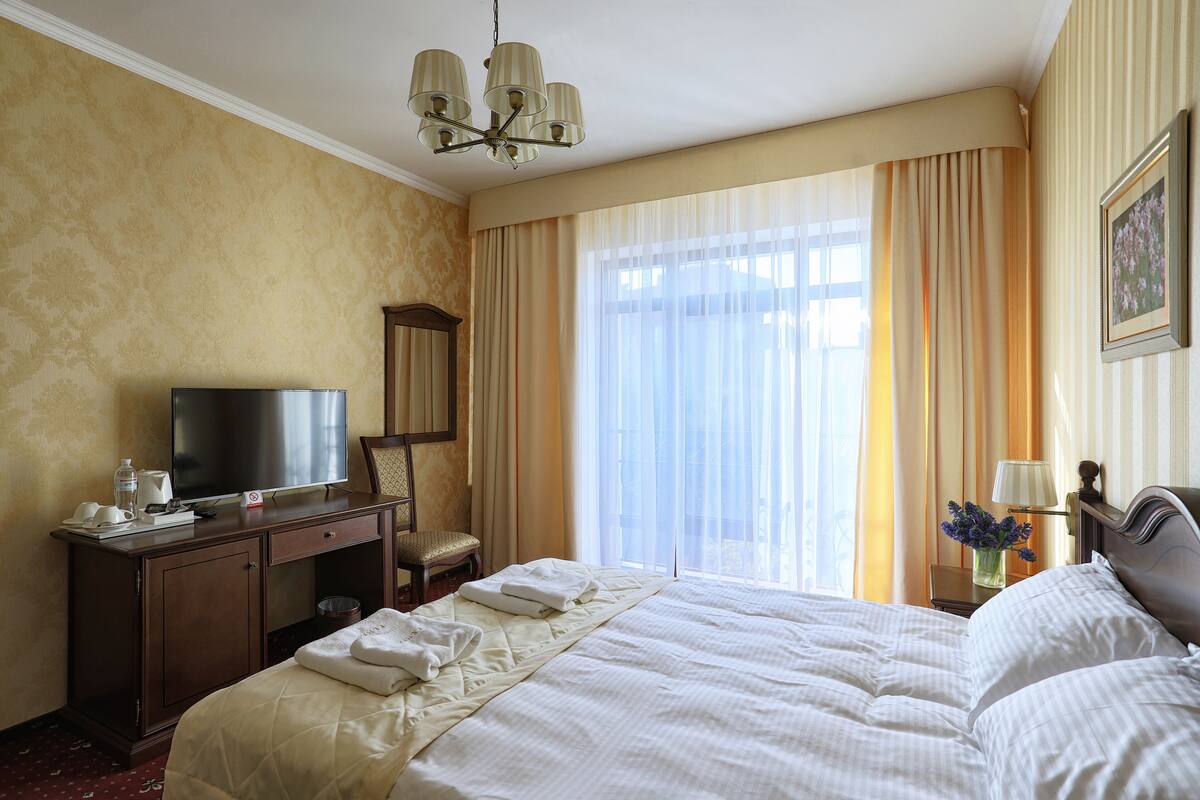A hotel room with a large bed, a desk with a television, a chair, and a nightstand with a lamp and flowers.