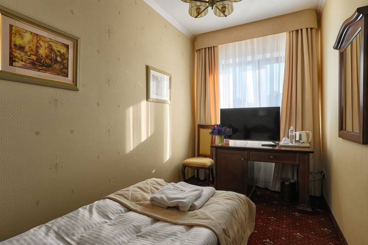 A hotel room with a bed, a desk with a chair, a television, and a window with curtains.