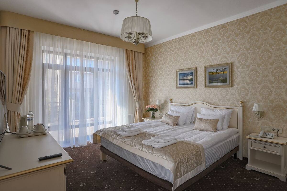 A hotel room with a large bed, a bedside table with a lamp, a desk with a telephone, and a window with curtains.
