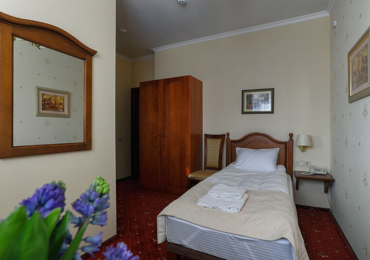 Una habitación de hotel con cama, un armario de madera, una silla, una mesita de noche con lámpara y un cuadro en la pared.