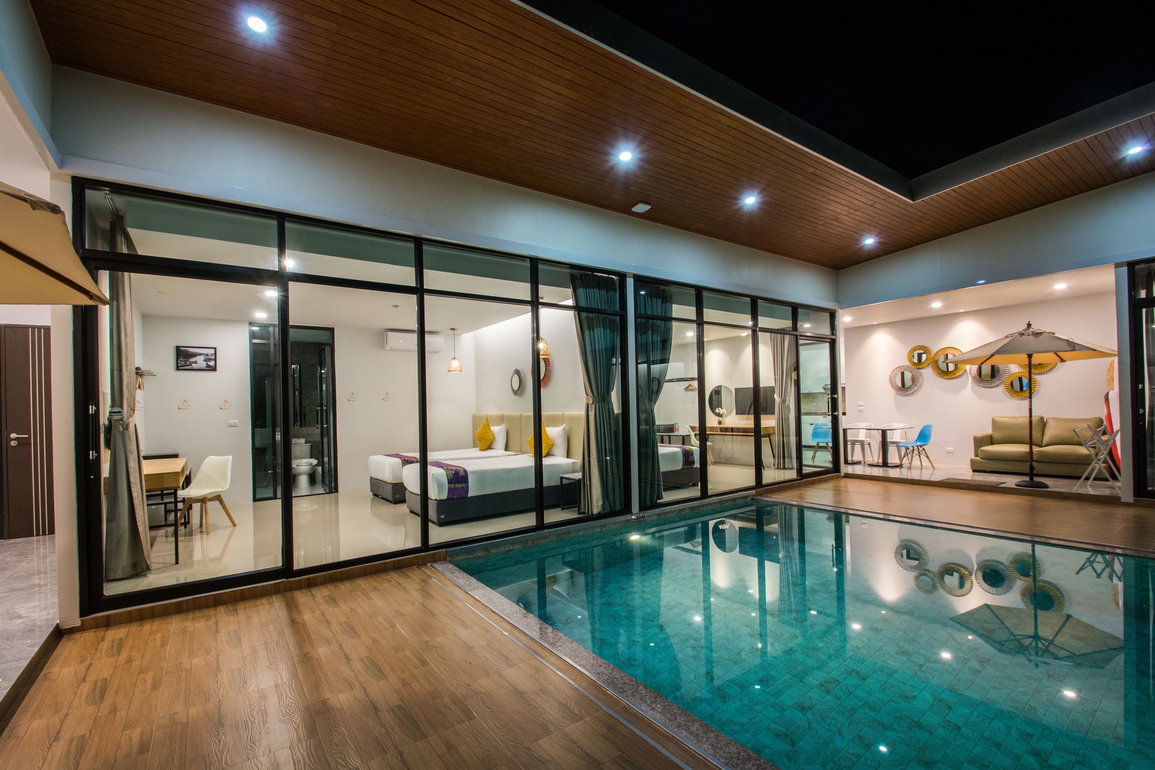 A modern indoor swimming pool with glass walls, wooden flooring, and a cozy seating area with a sofa and chairs.