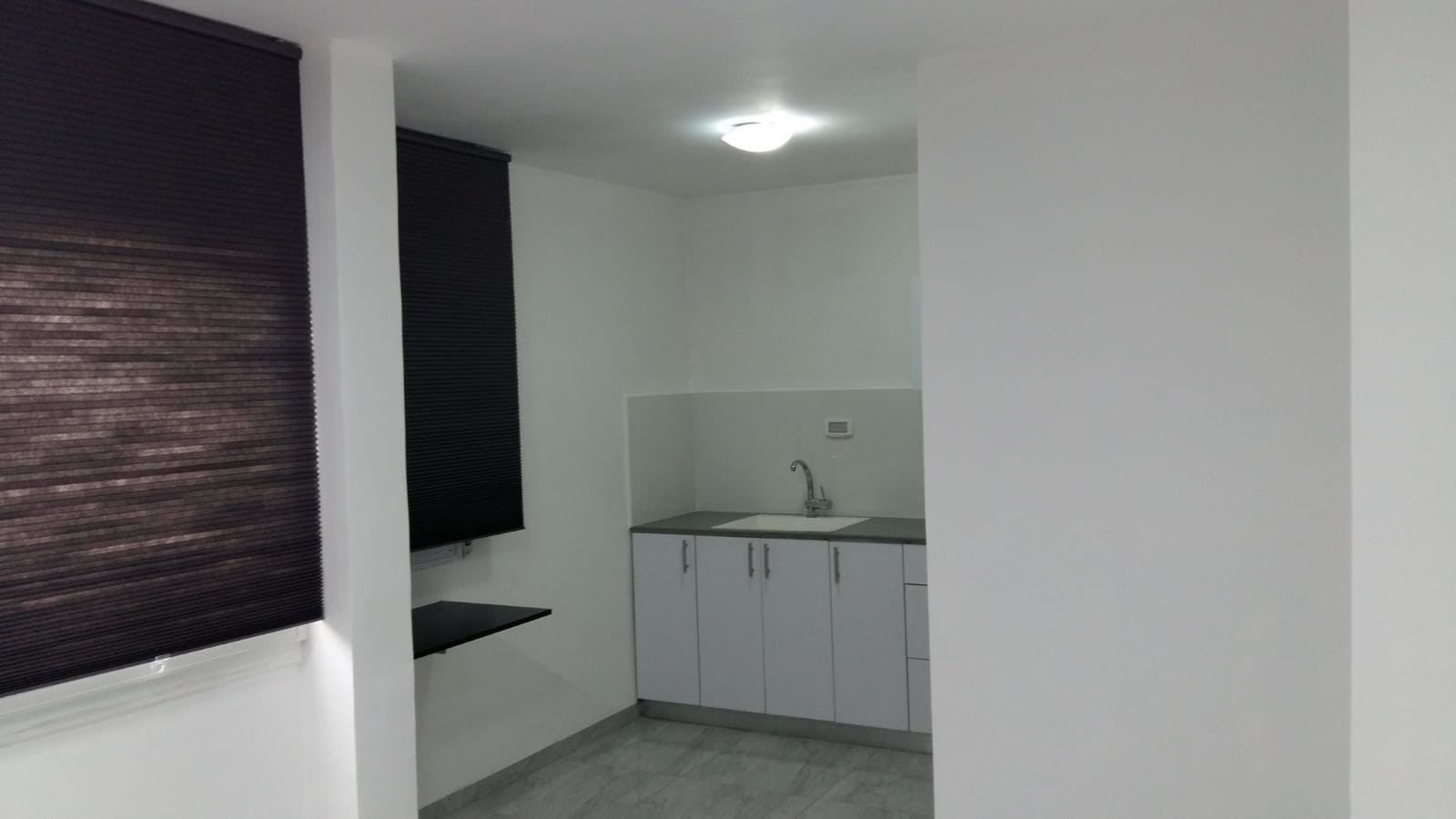 A small, compact kitchen with a single sink, white cabinets, and a dark-colored window blind.