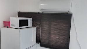 Una kitchenette compacta con microondas y refrigerador, una pequeña cesta rosa y un ventanal con persianas.