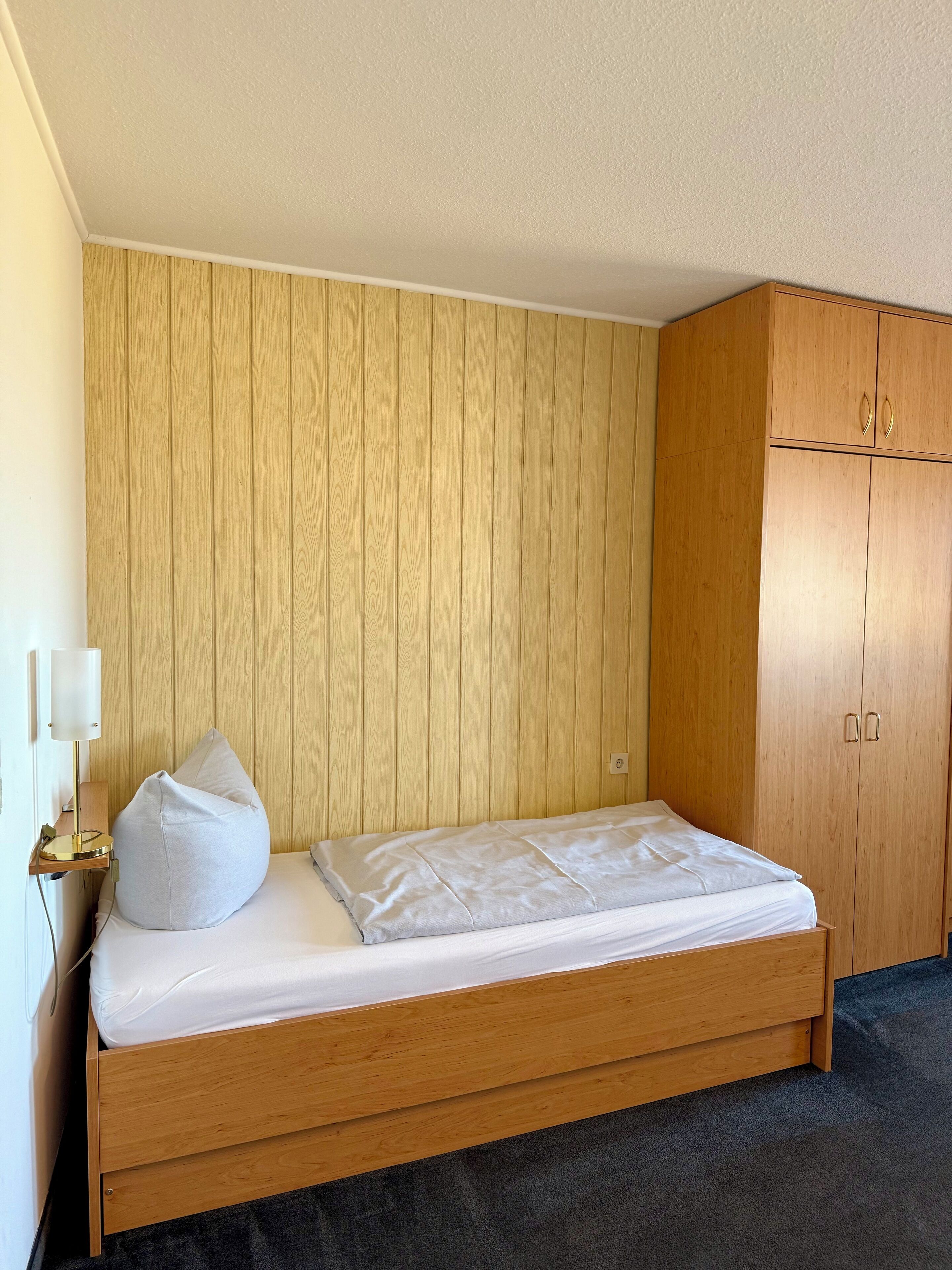 Una cama individual con estructura y cabecero de madera, frente a una pared de madera. Una lámpara de noche y una almohada blanca están sobre la cama. Un armario de madera con dos puertas se encuentra a la derecha.