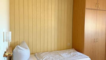 Un lit simple avec un cadre en bois et une tête de lit adossée à un mur en bois. Une lampe de chevet et un oreiller blanc sont posés sur le lit. Une armoire en bois à deux portes se trouve à droite.