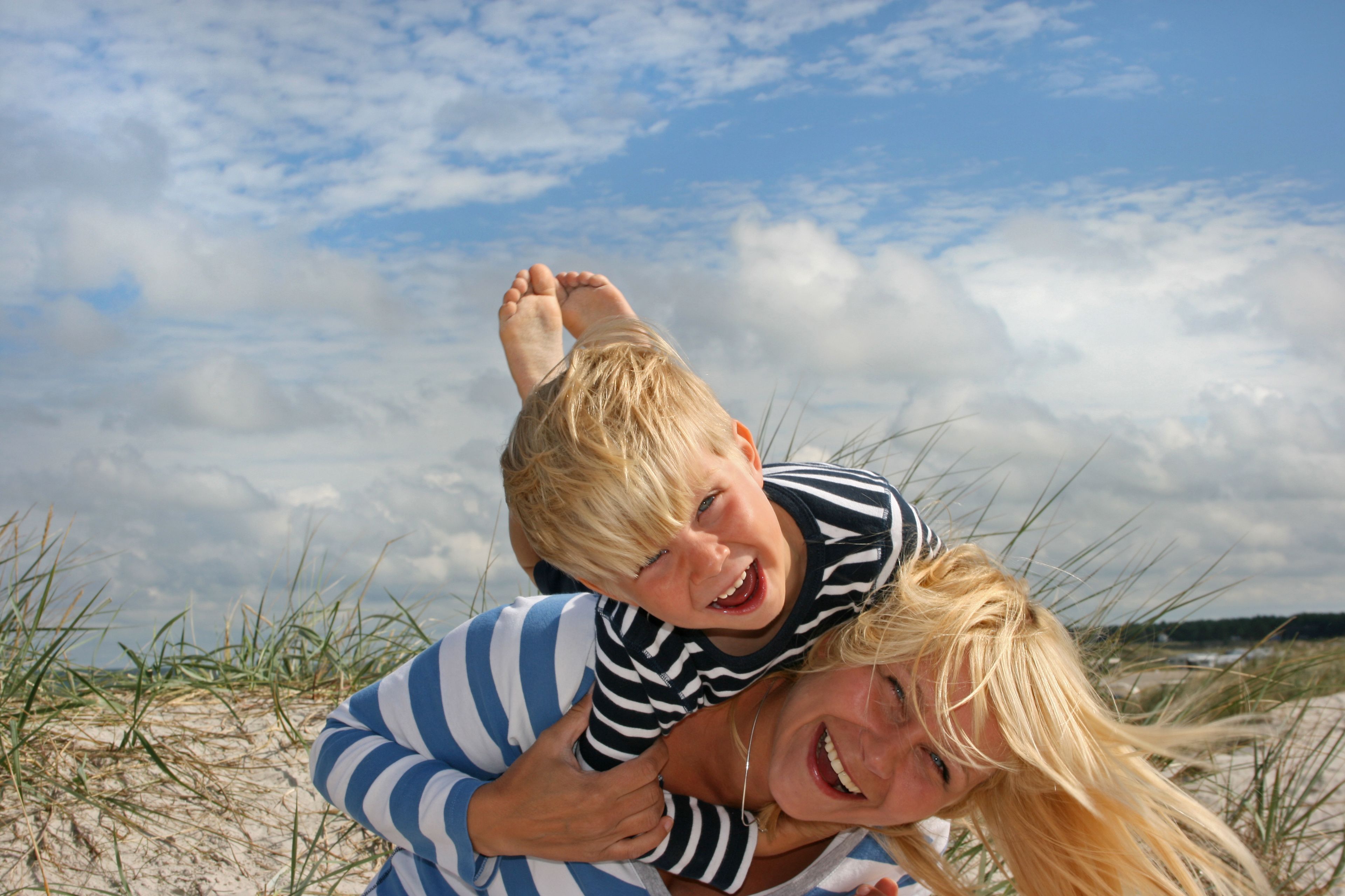 A person carrying another on their back, with a sandy beach and dunes in the background.