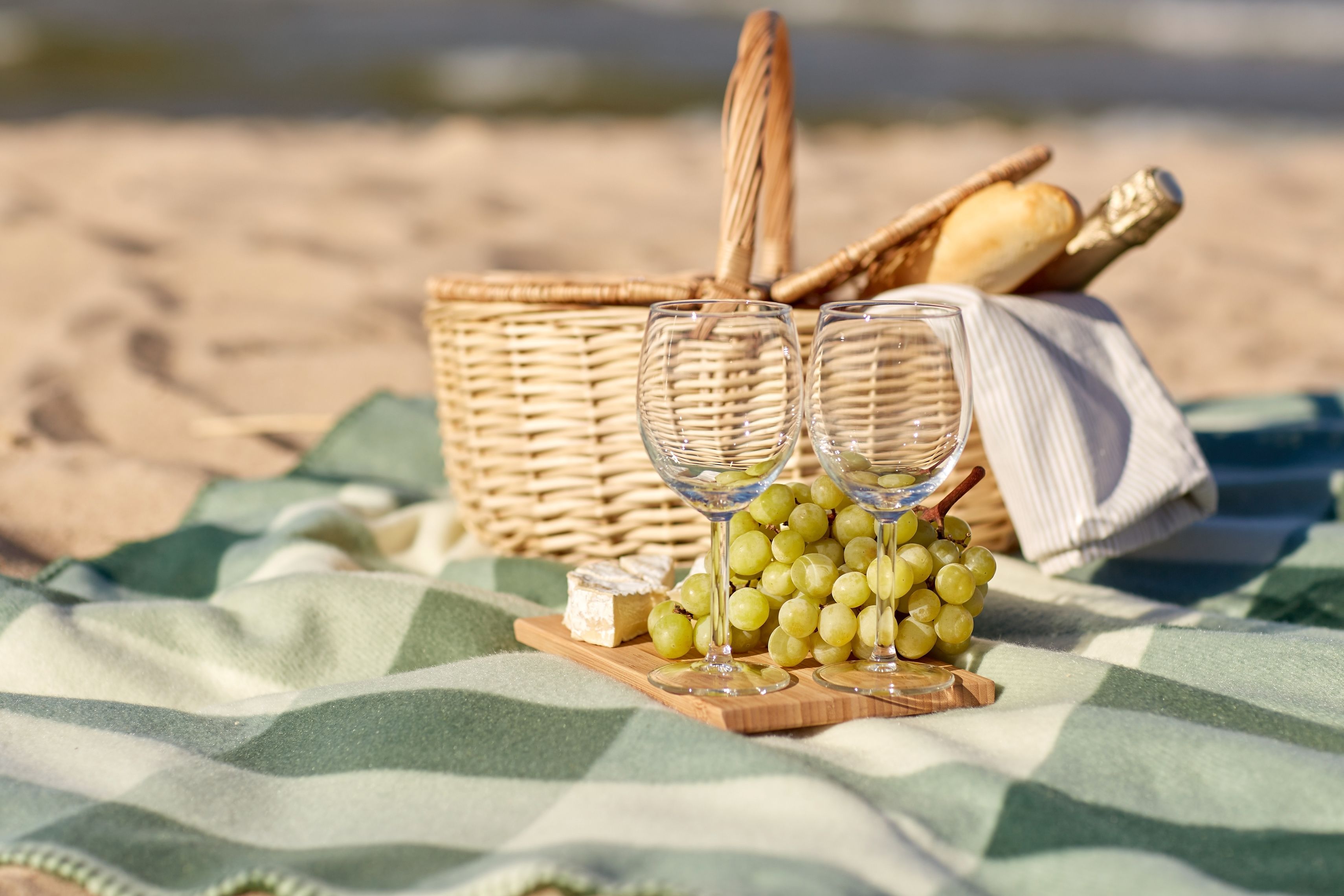 Un montaje para picnic en la playa con una cesta de mimbre, barras de pan, tela, una tabla de madera con uvas y queso, y dos copas de vino.