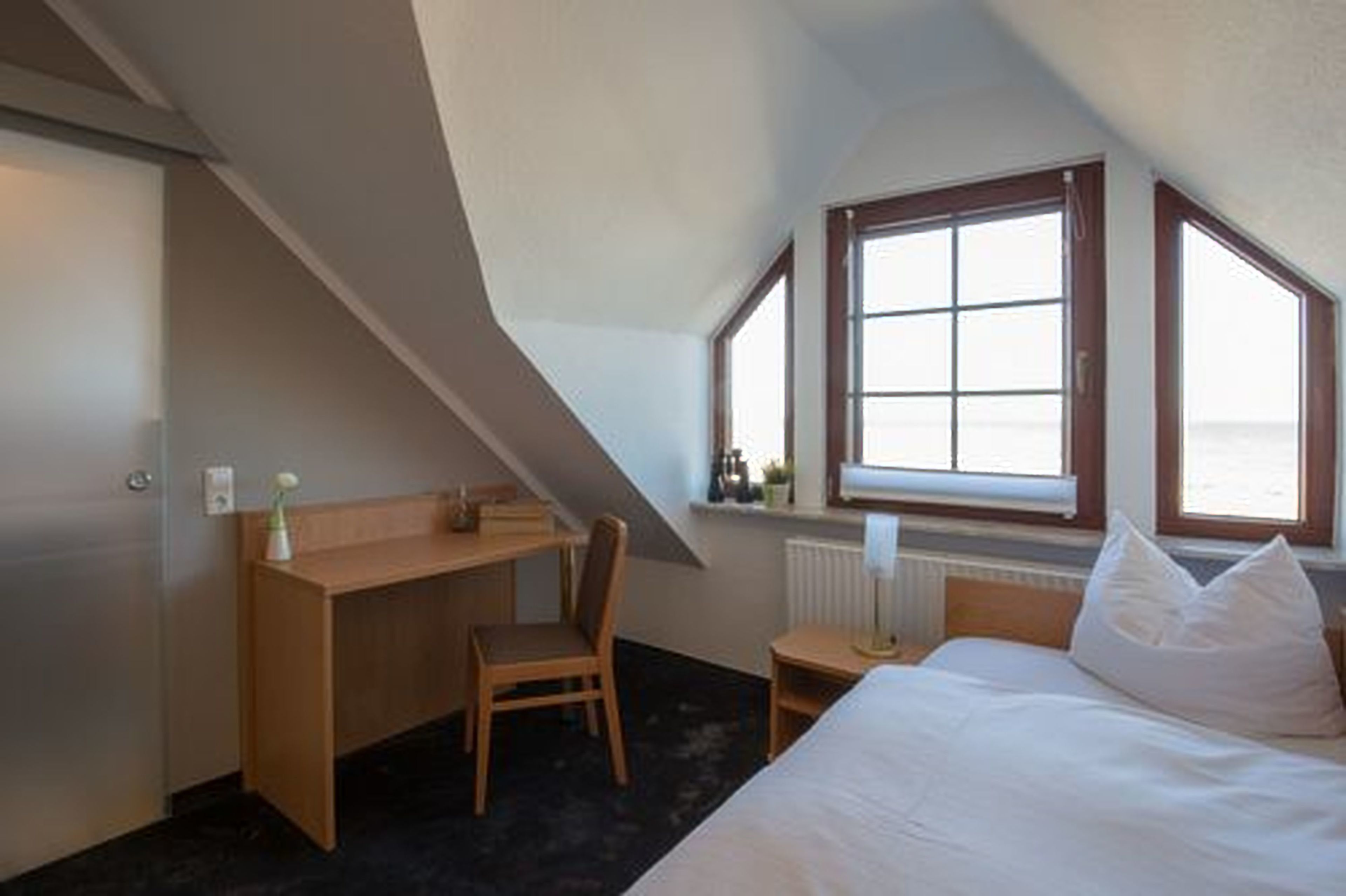 Ein Schlafzimmer mit schrägverdeckter Decke, einem Holztisch, einem Stuhl, einem Bett mit weißer Bettwäsche und zwei Fenstern.