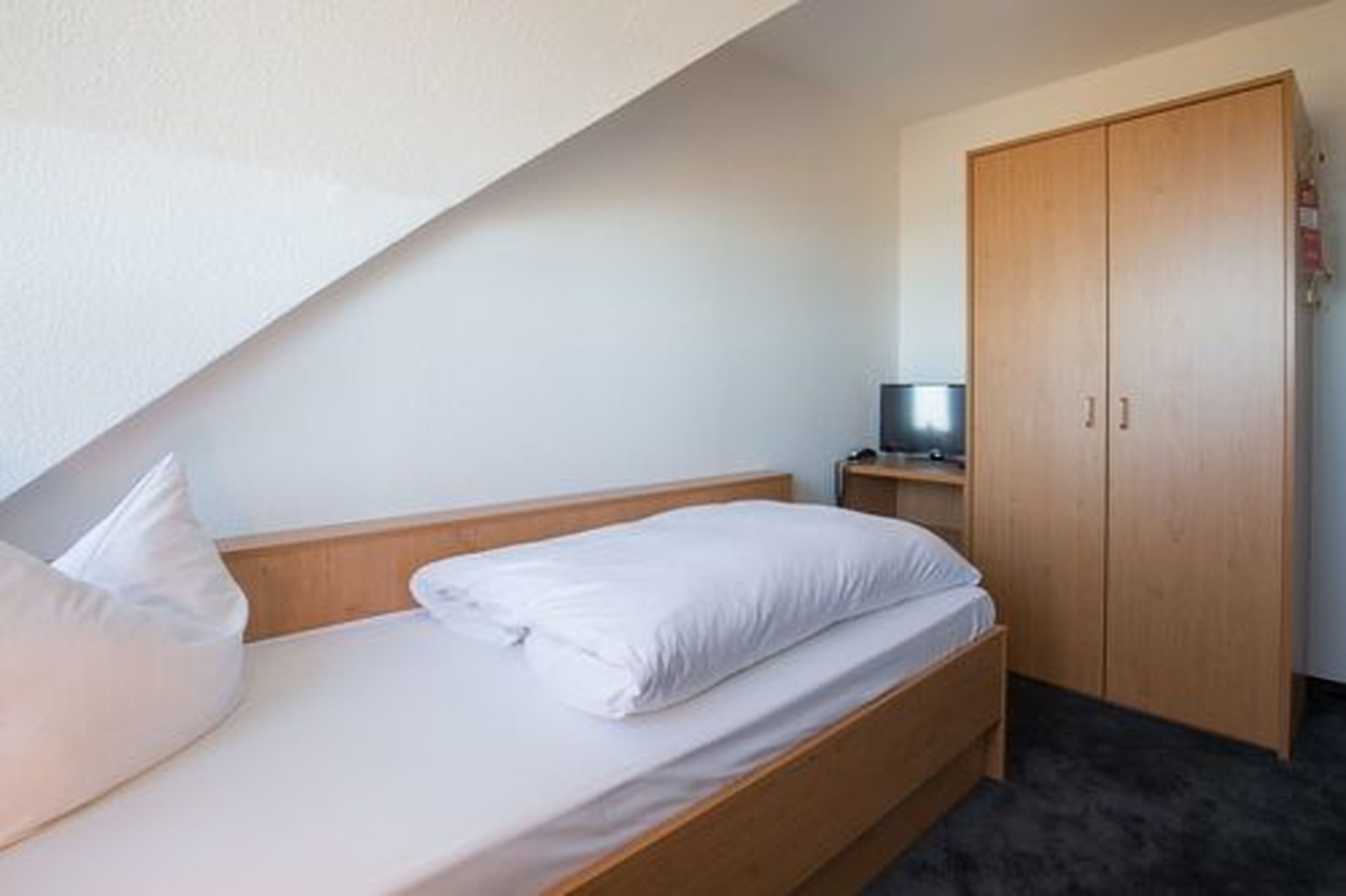 Ein Einzelbett mit weißer Bettwäsche in einem Zimmer, das einen Holzschrank und einen kleinen Tisch mit einem Fernseher enthält.