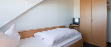Ein Einzelbett mit weißer Bettwäsche in einem Zimmer, das einen Holzschrank und einen kleinen Tisch mit einem Fernseher enthält.
