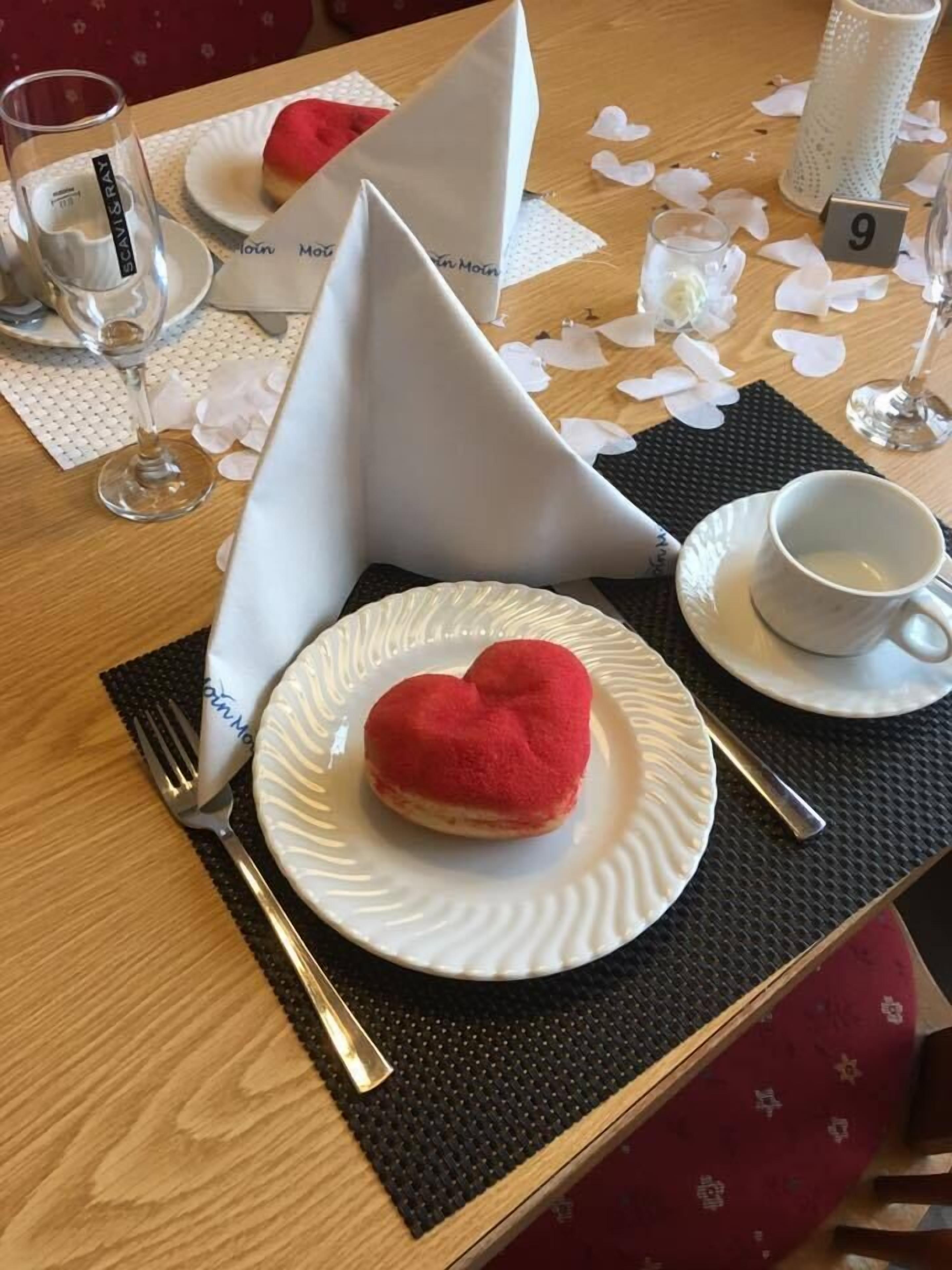 Ein Tisch, gedeckt mit einer herzförmigen Patisserie auf einem weißen Teller, einem Glas Wasser, einer Tasse und einer Serviette mit einem Logo.