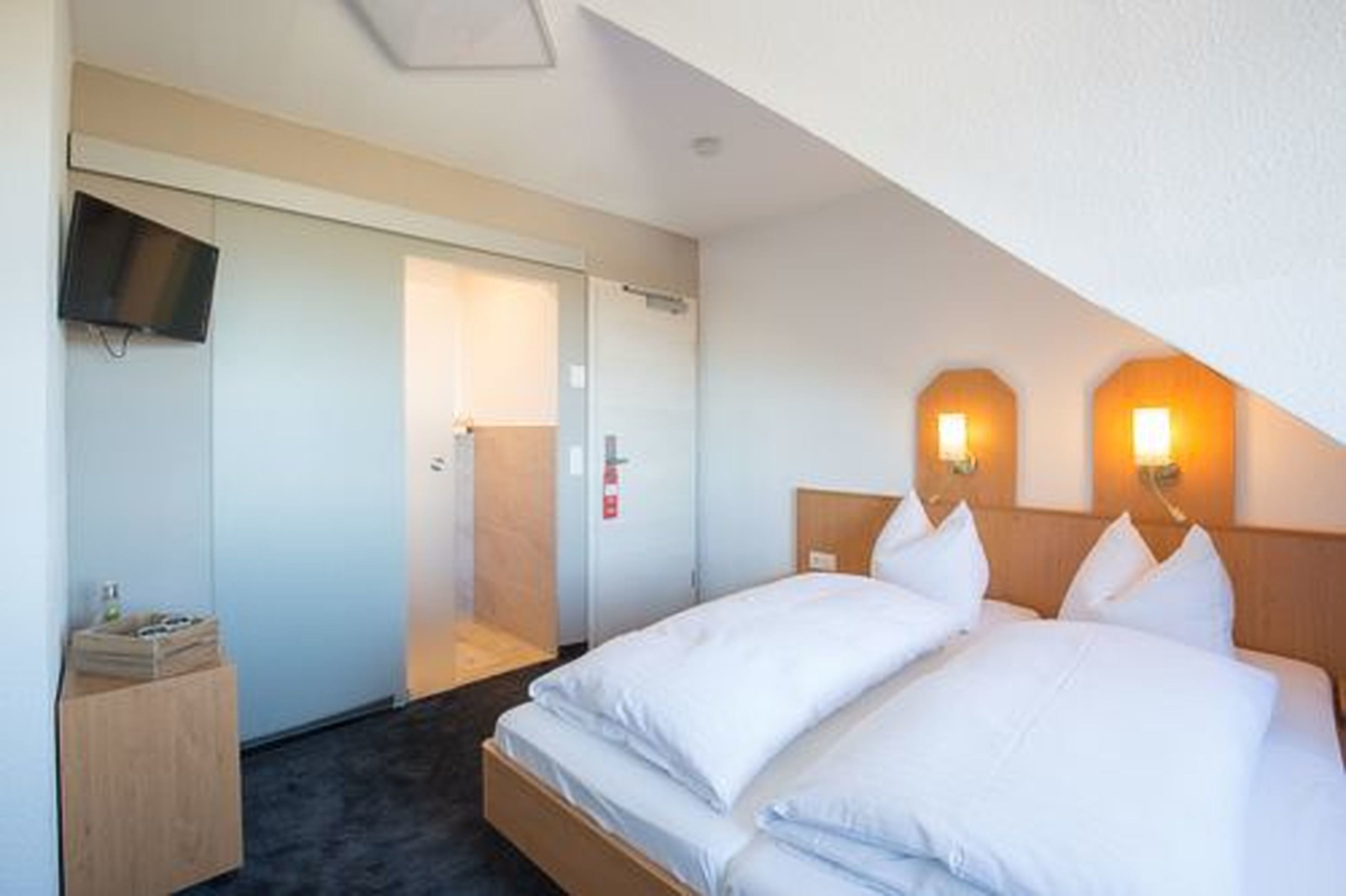 A hotel room with a bed, a TV mounted on the wall, and a wooden nightstand.