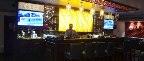 A bar area with a bartender, bar stools, and two TVs mounted on the wall.