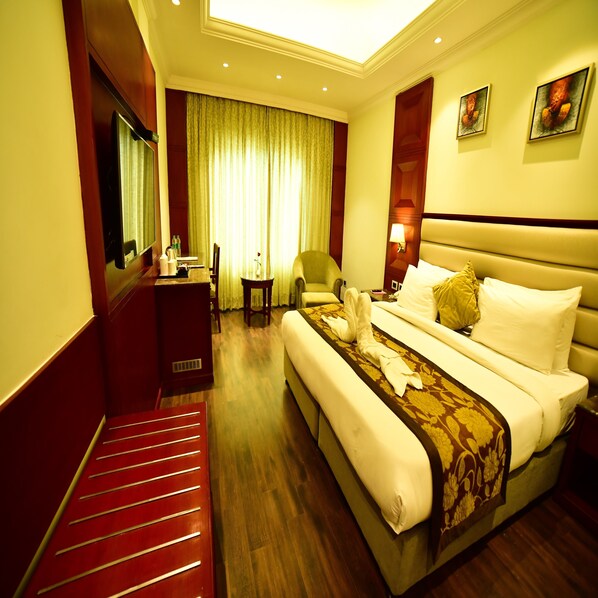 A hotel room with a large bed, a desk, a chair, a small table, and a mirror. - Hotel Milan Palace (Prayagraj)