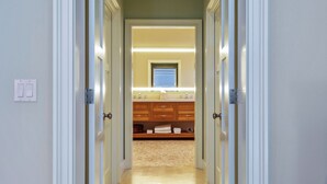 Un couloir menant à une salle de bain équipée d’un meuble-lavabo en bois, d’un miroir et d’un porte-serviettes.