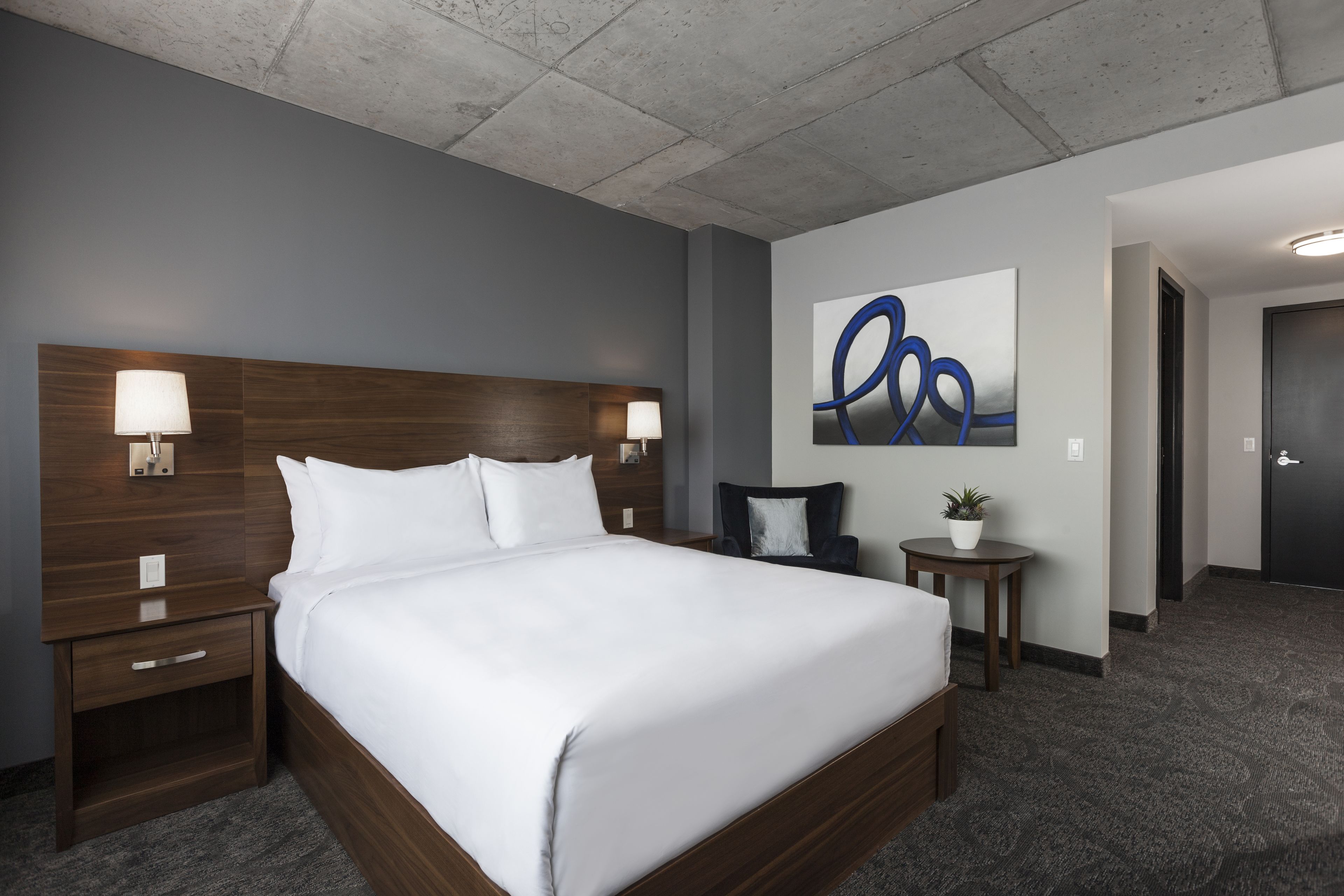 A hotel room with a large bed, two bedside tables with lamps, a small side table with a plant, and a modern abstract painting on the wall.