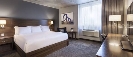 A hotel room with a large bed, bedside tables, a desk, a chair, a potted plant, an air conditioning unit, and a window with curtains.