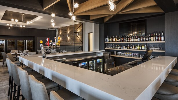 Zona de bar moderna con una barra alargada, varios taburetes y una amplia selección de licores.