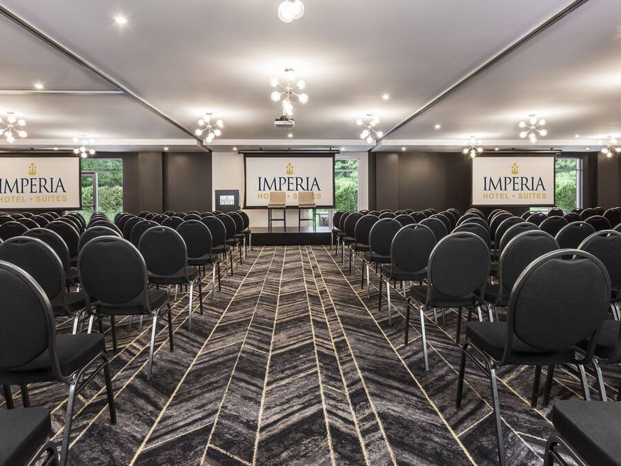 A conference room with rows of chairs, a stage, and banners reading "IMPERIA HOTEL SUITES".