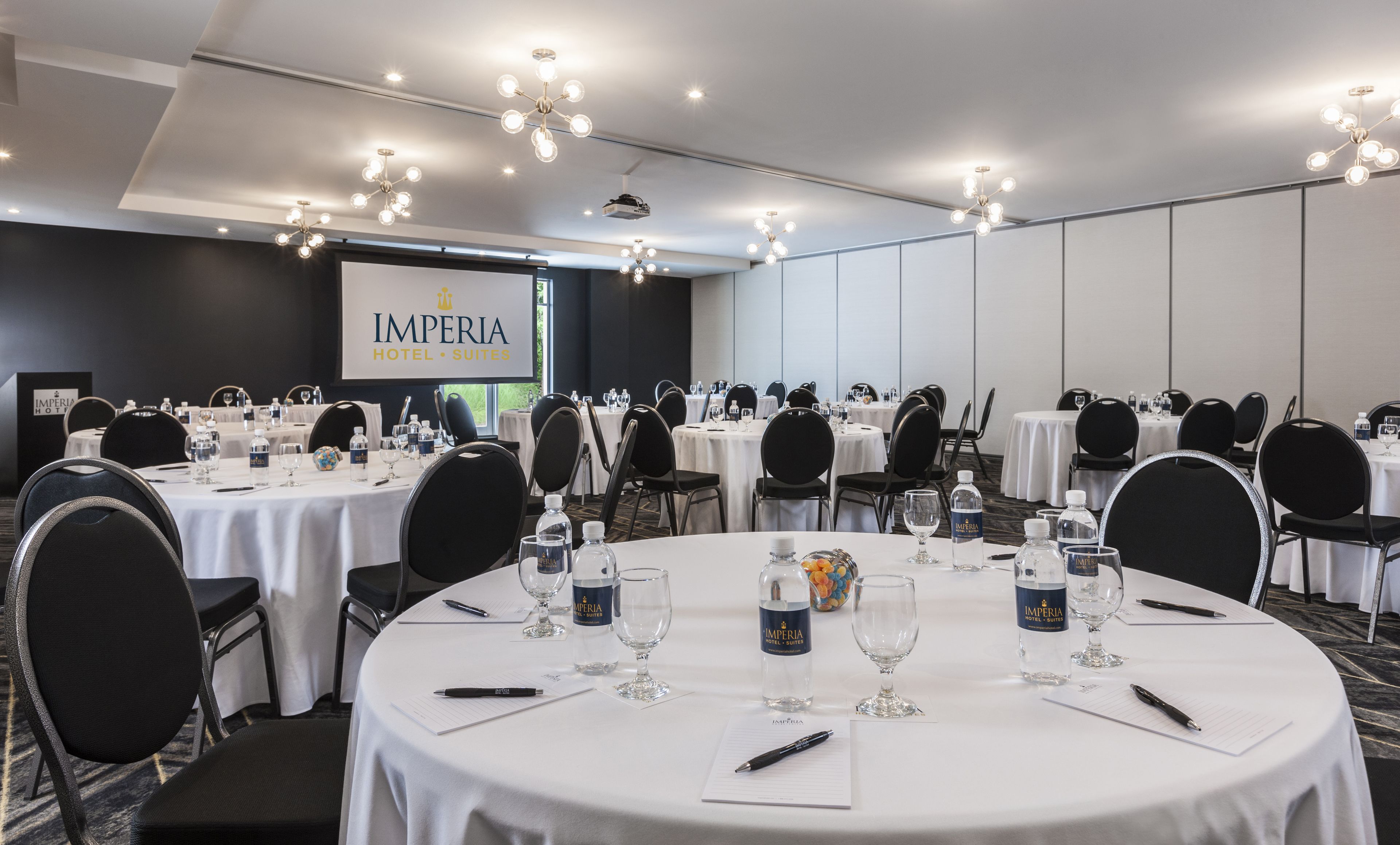 A conference room at the Imperia Hotel Suites with round tables, chairs, and water bottles.