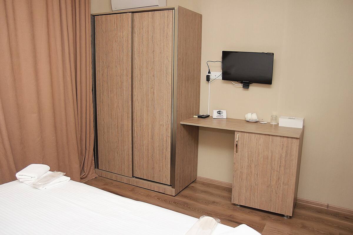 A hotel room with a bed, a wardrobe, a desk with a flat-screen TV, and a bedside table.