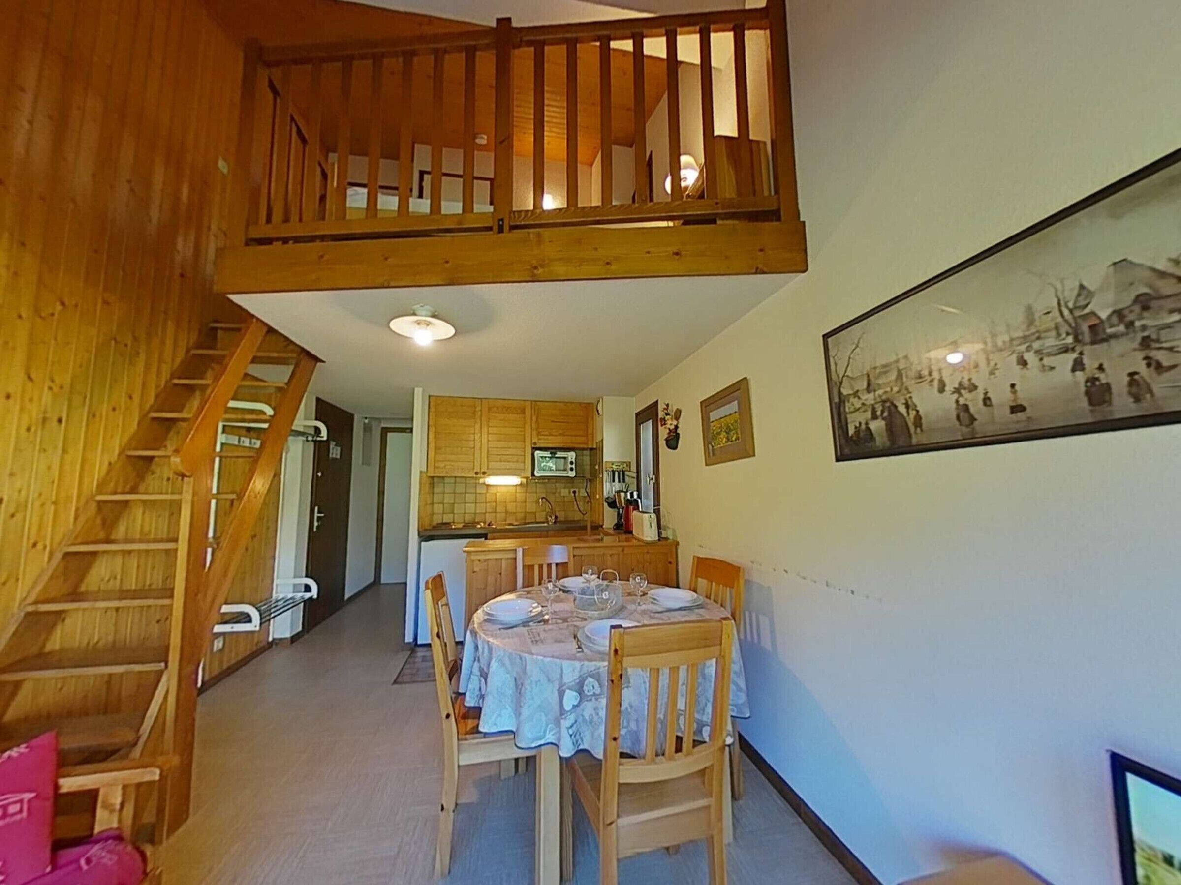 A dining area with a wooden table set for four, a kitchen in the background, and a staircase leading to an upper level.
