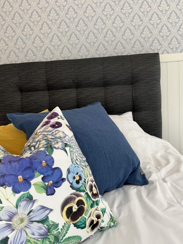 Una cama con cabecera oscura, ropa de cama blanca y almohadas decorativas con motivos florales.