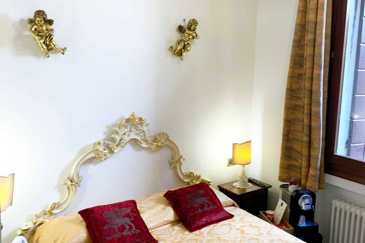 A hotel room with a bed, a chandelier, a nightstand with a lamp, and a window with curtains.