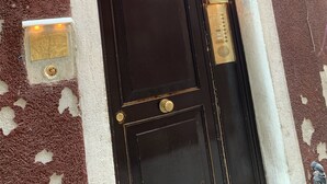 Une porte noire, dotée d’une poignée et d’un heurtoir en laiton, encadrée de pierre blanche et placée au-dessus d’un mur rouge à la texture particulière.