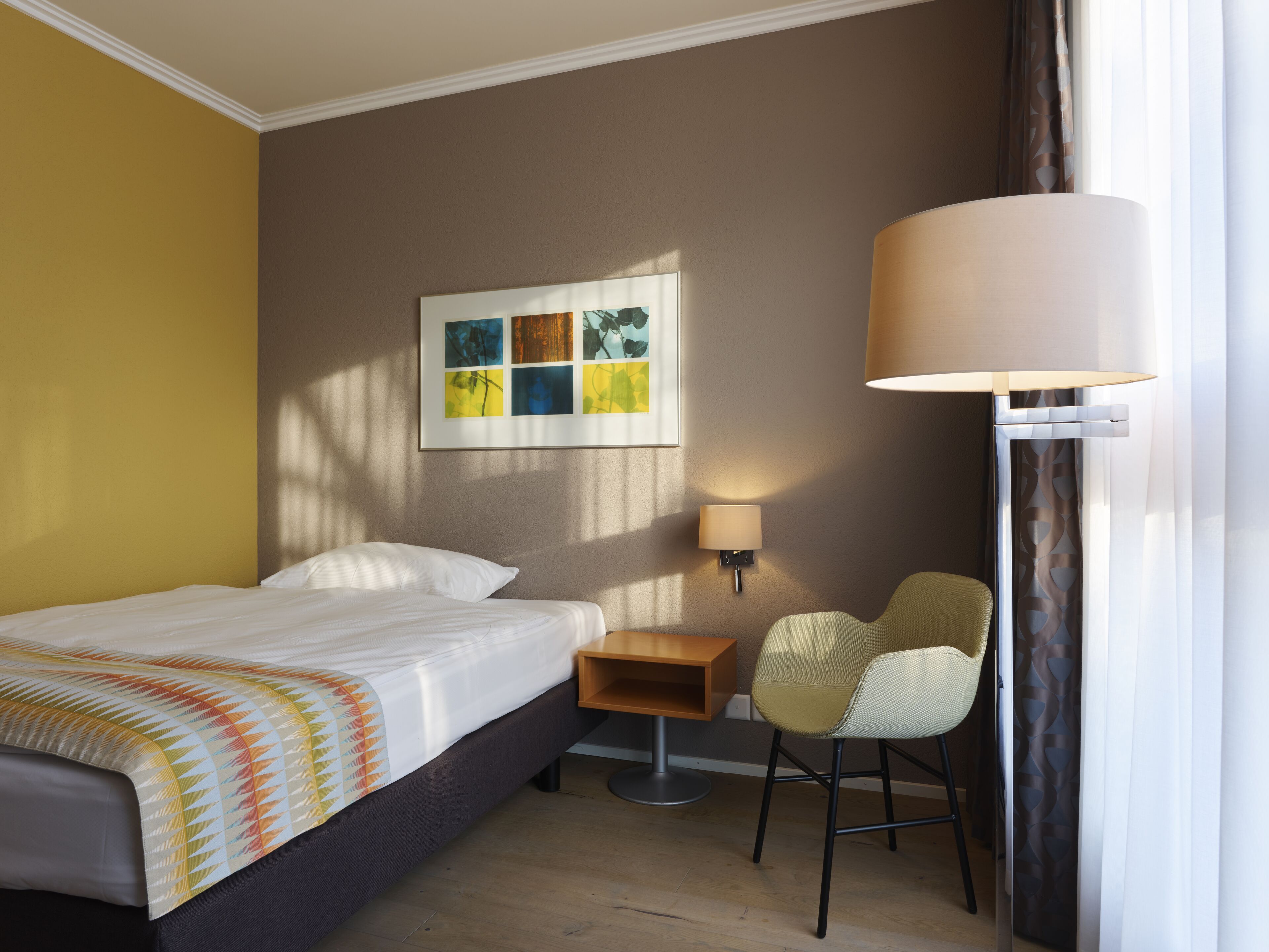 A hotel room with a bed, a nightstand, a chair, a lamp, and a painting on the wall.
