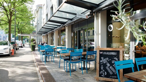 An outdoor café with blue chairs and tables, a chalkboard menu, and a sign welcoming visitors. - Hotel Berchtold (Burgdorf)