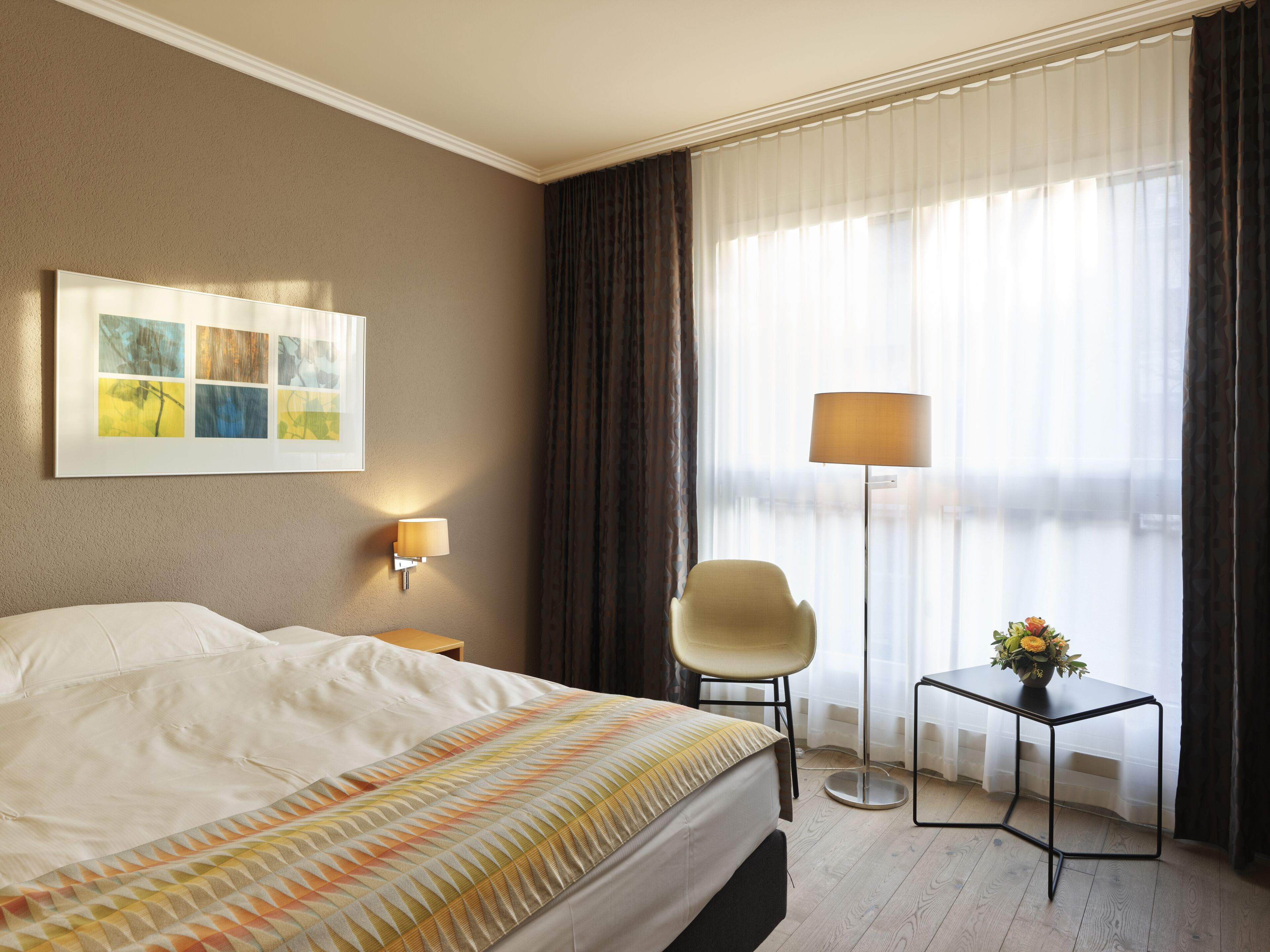 Habitación de hotel con cama, una silla, una mesita con un jarrón de flores, una lámpara y un cuadro en la pared.