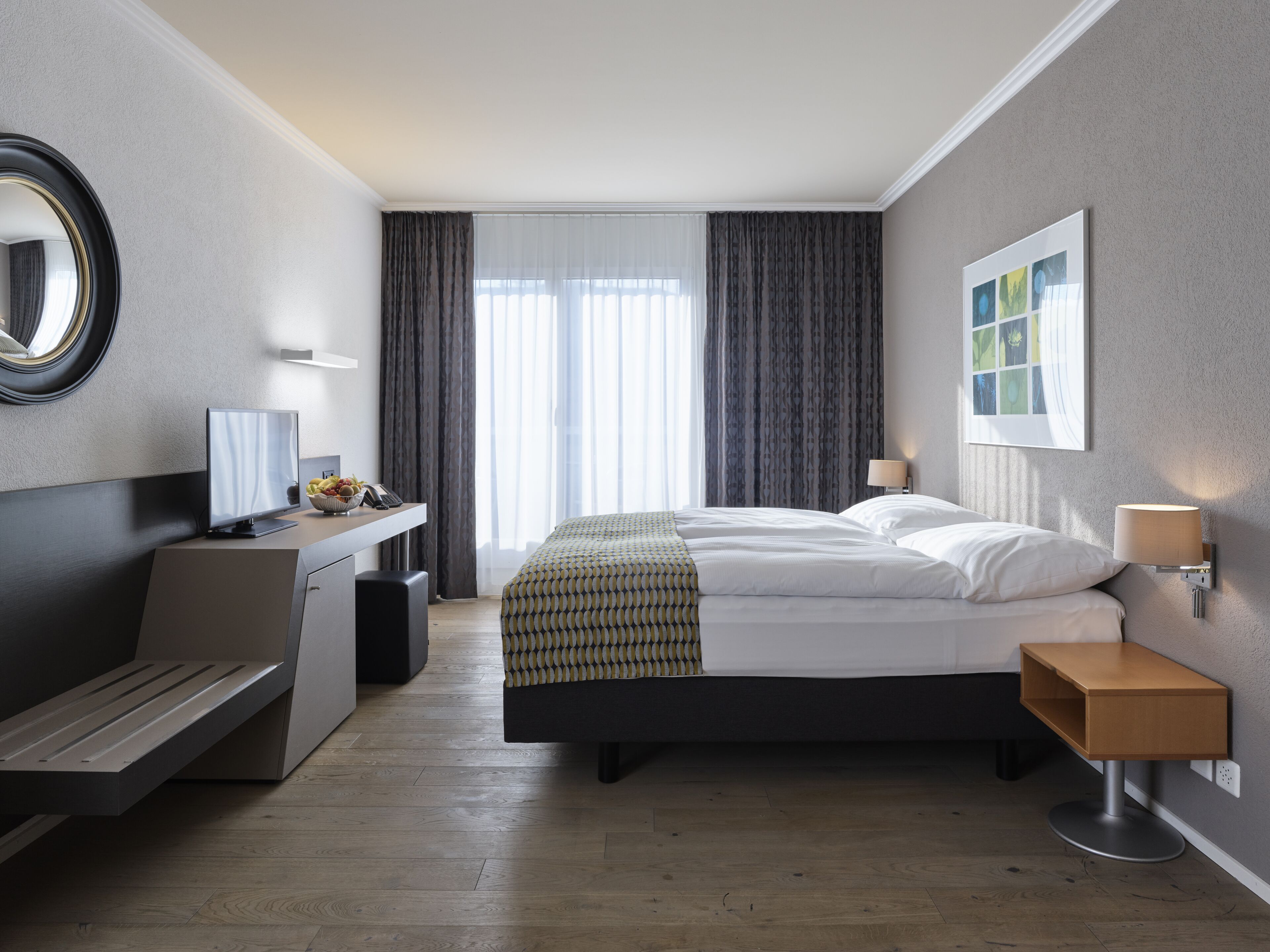 A modern hotel room with a bed, bedside table, desk, and a large window with curtains.