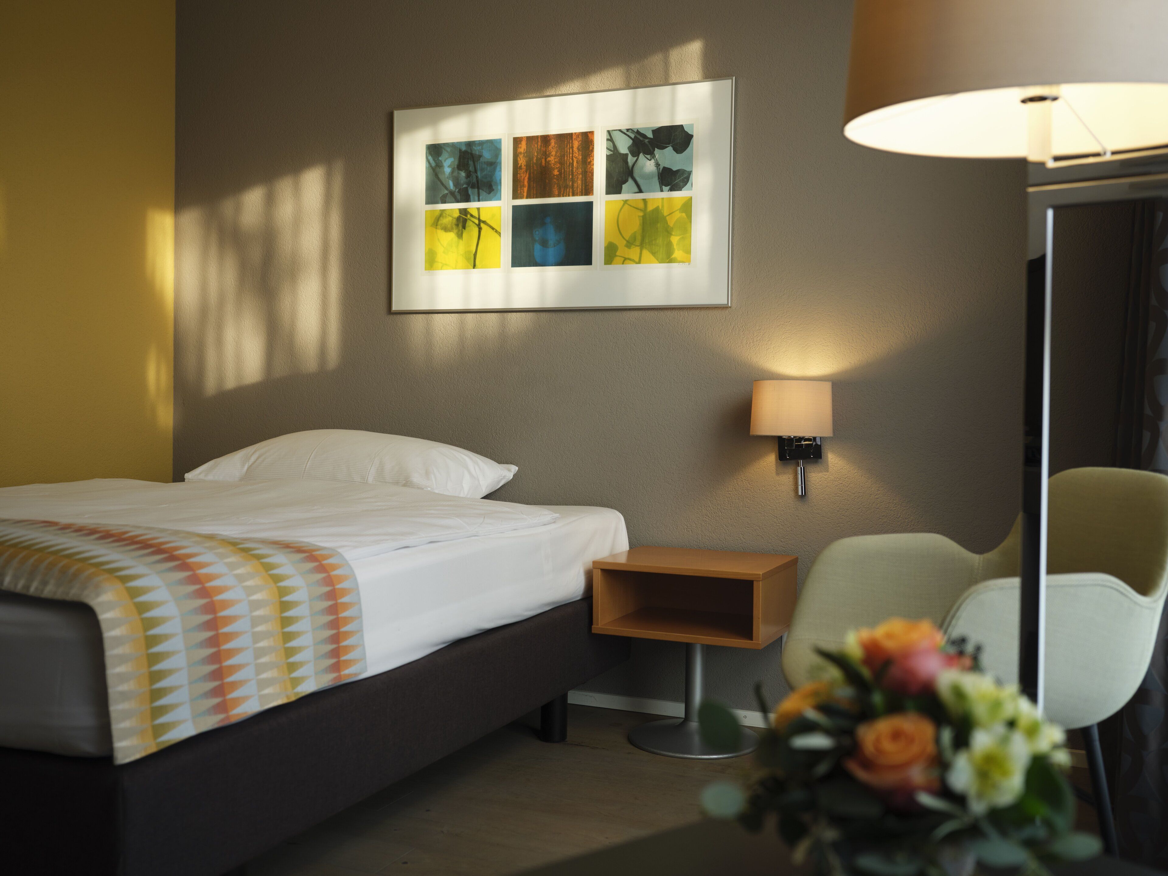 Ein Hotelzimmer mit einem Bett, einem Nachttisch, einem Sessel, einer Wandlampe und einem Bildrahmen über dem Bett.