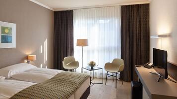 Une chambre d’hôtel avec un lit, deux chaises, un bureau avec un ordinateur et une fenêtre avec des rideaux.