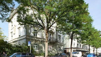 Un edificio de estilo arquitectónico clásico, con un árbol grande en primer plano, un letrero de estacionamiento y un cartel azul que indica un hotel.