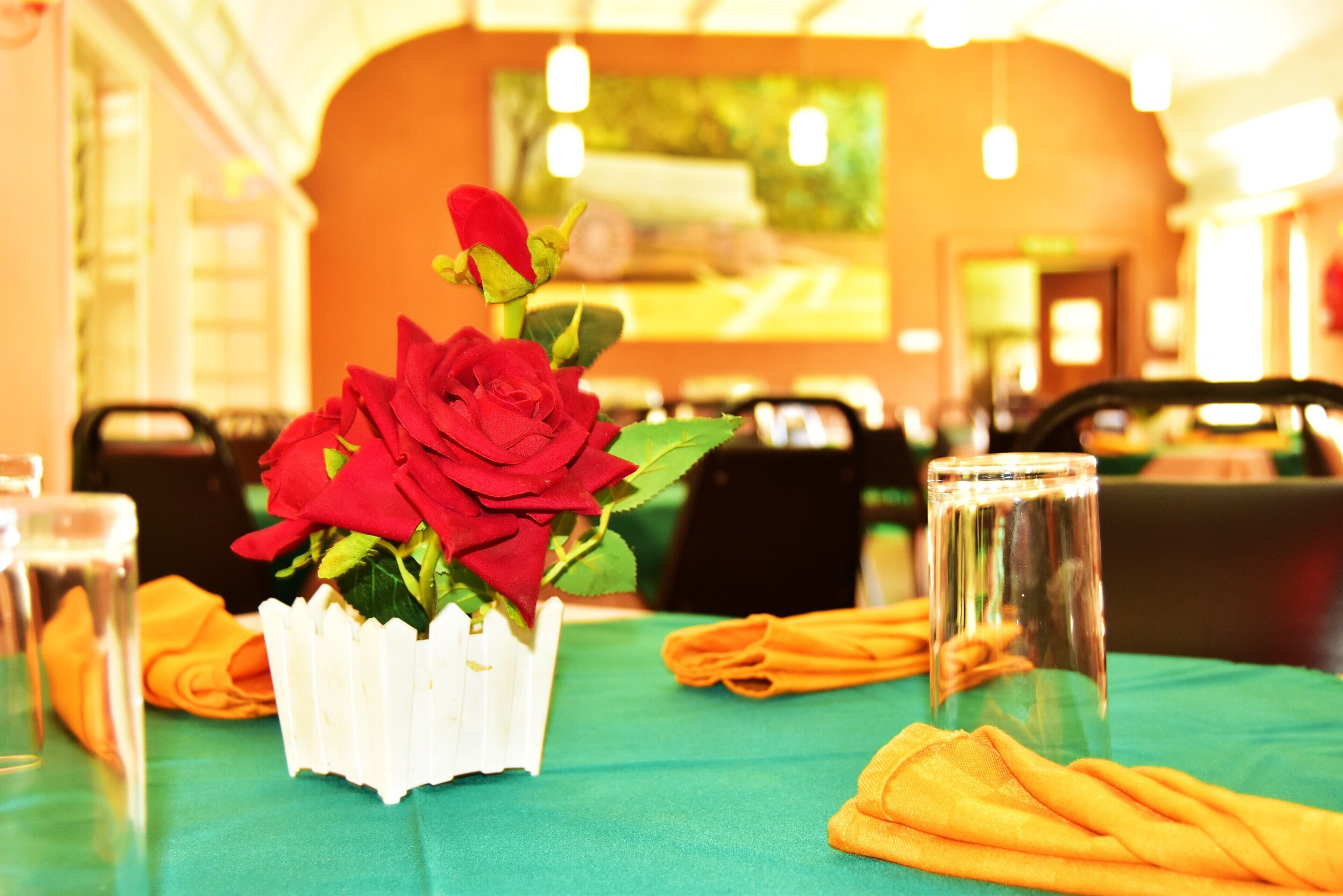 Une table à manger ornée d’un vase blanc contenant des roses rouges, d’un verre transparent et de serviettes orange.