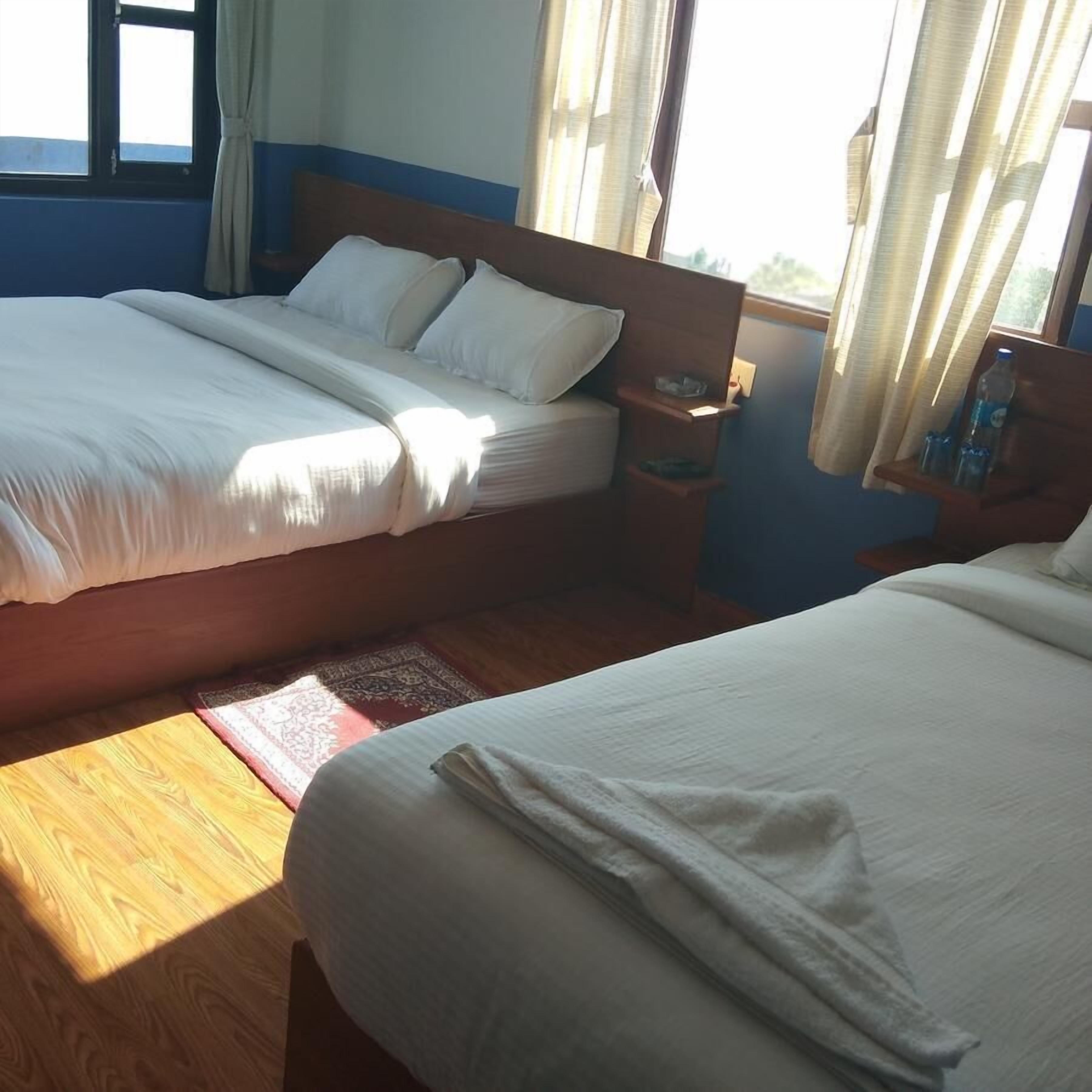 A hotel room with two beds, wooden flooring, a nightstand, and a window with curtains.
