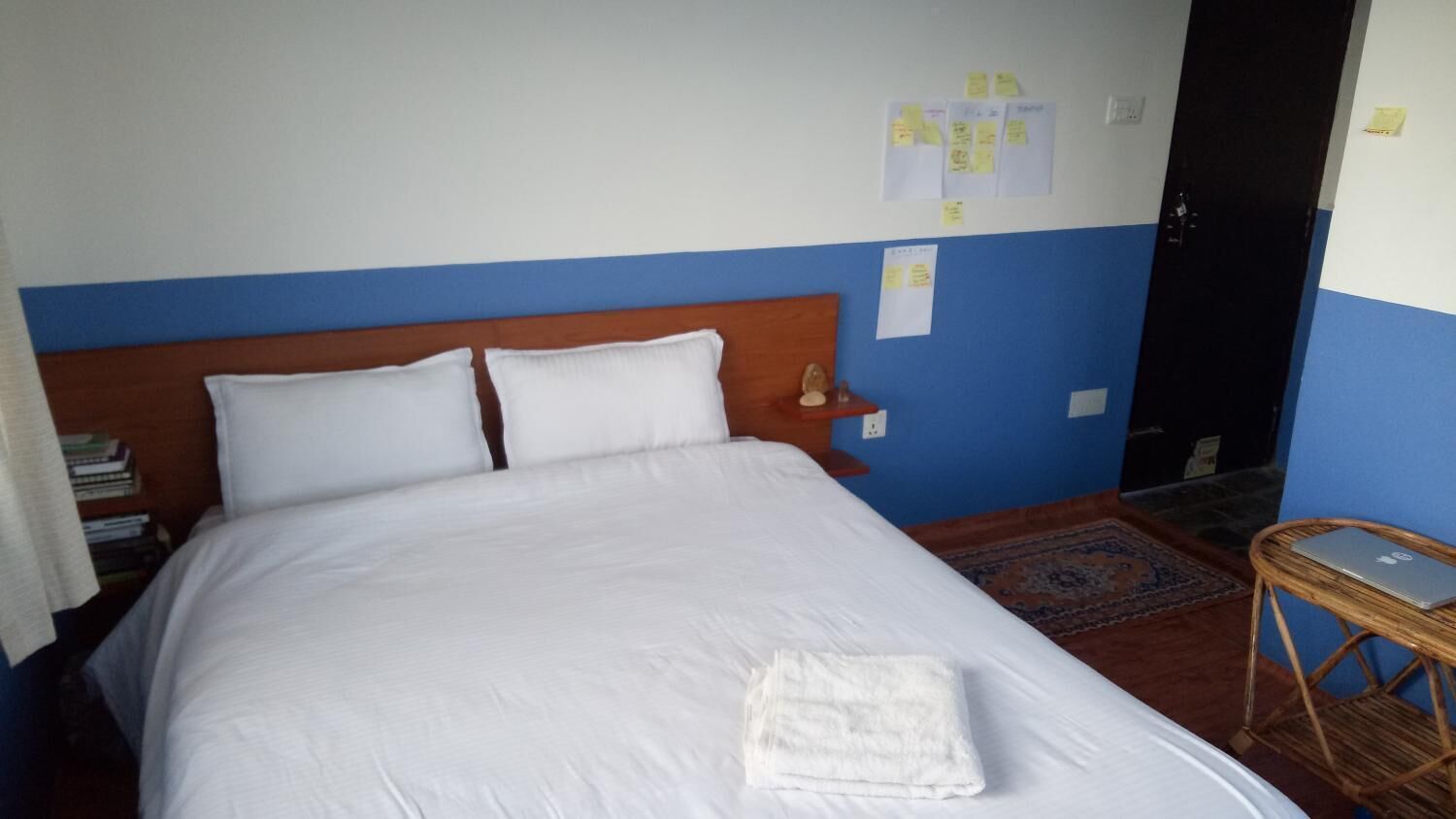 Una cama bien tendida con sábanas blancas, un cabecero de madera y una mesita de noche con una lámpara. Hay notas sujetas a la pared y una silla de mimbre con un ordenador portátil encima.