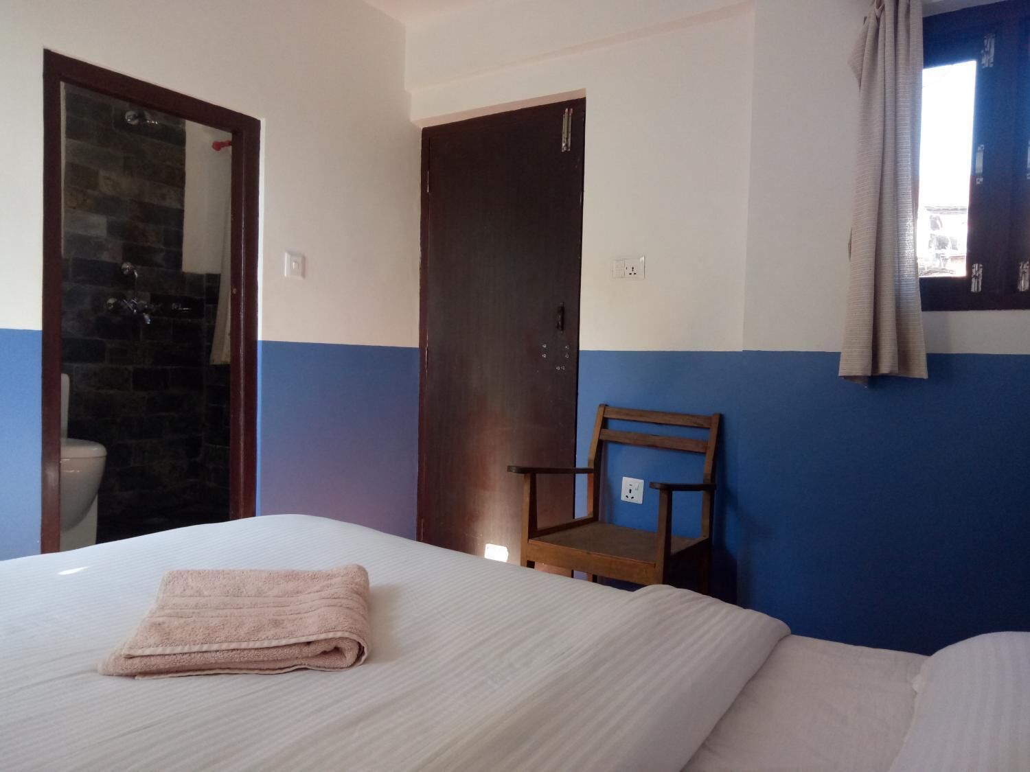 Quarto de hotel com cama, uma cadeira de madeira, banheiro com chuveiro e porta.