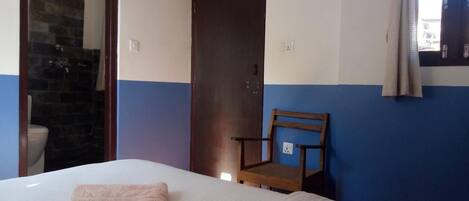 Quarto de hotel com cama, uma cadeira de madeira, banheiro com chuveiro e porta.