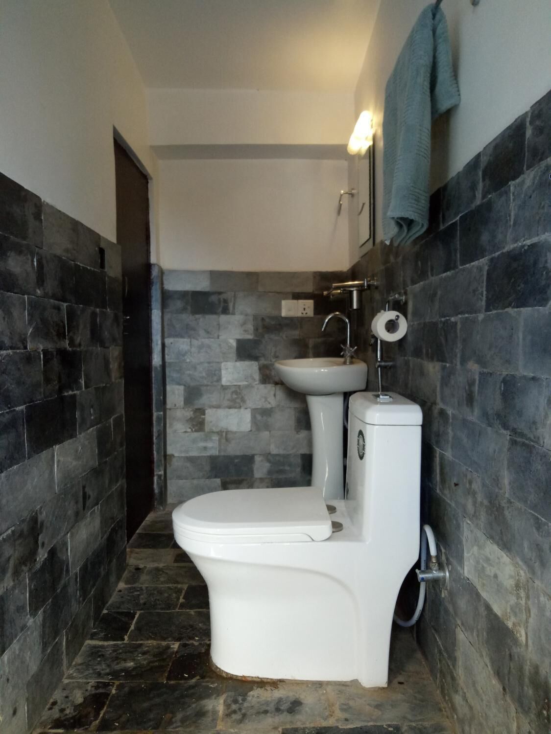 A bathroom with a white toilet, a sink, and a shower. The walls are tiled with dark stones.
