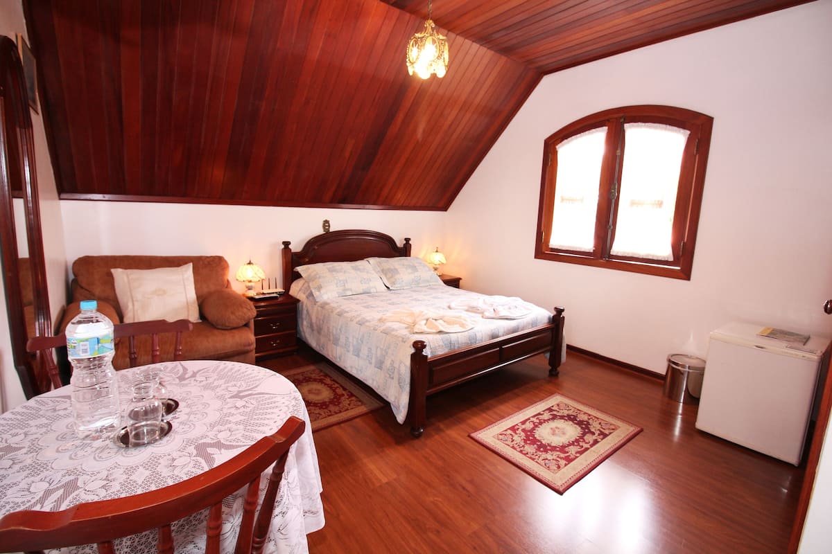 Ein Schlafzimmer mit Holzbalkendecke, einem Bett mit Kopfteil, einem Nachttisch, einem Tisch mit Stuhl, einem Fenster und einem Kühlschrank.
