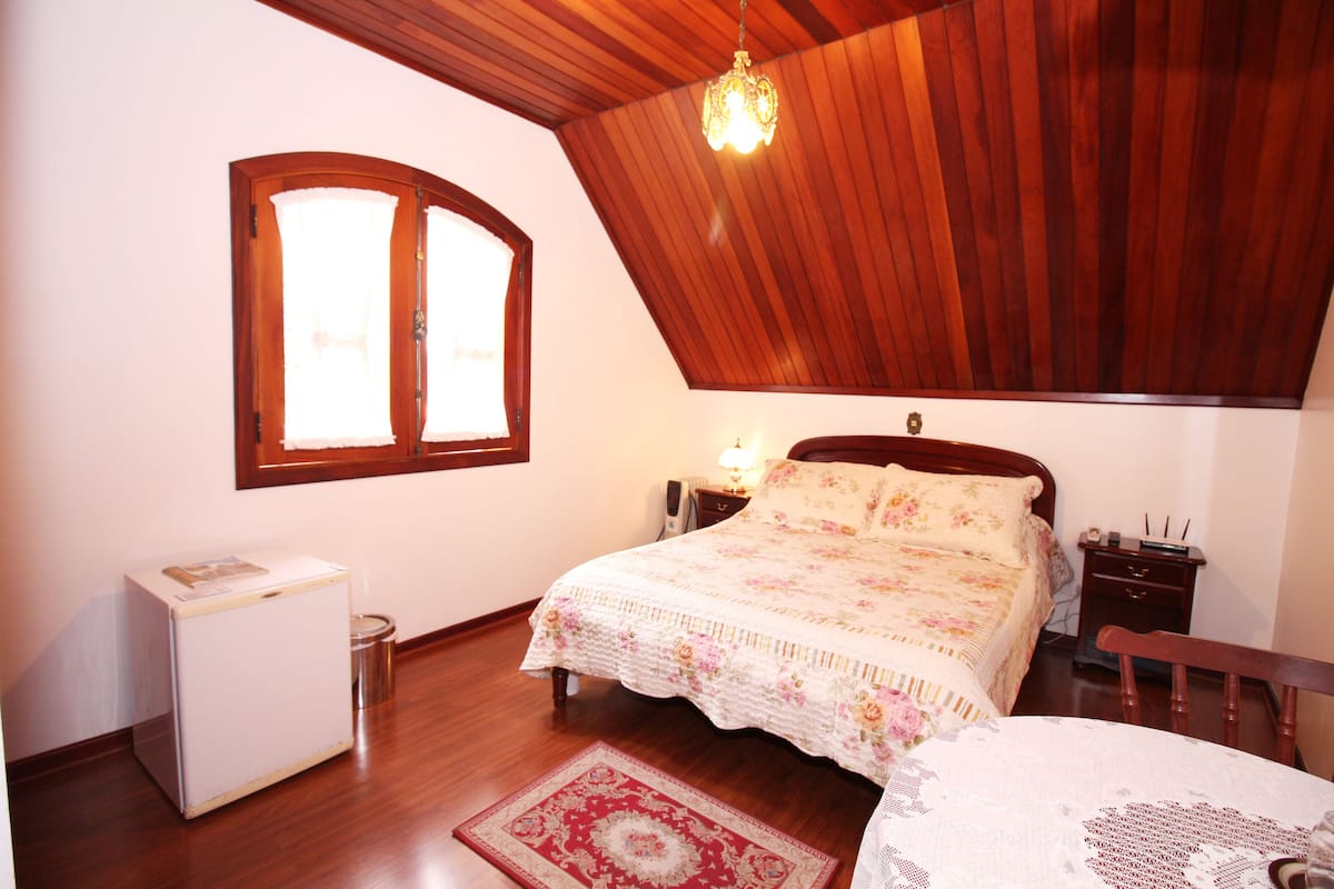 Ein Schlafzimmer mit Holzbalkendecke, einem Bett mit Blumenmuster-Bettwäsche, einem Nachttisch, einer kleinen weißen Kommode und einem Fenster mit Vorhängen.