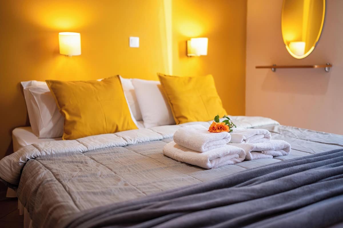 Un lit bien fait, avec des oreillers blancs et jaunes, une serviette pliée et une petite fleur posée sur le lit.