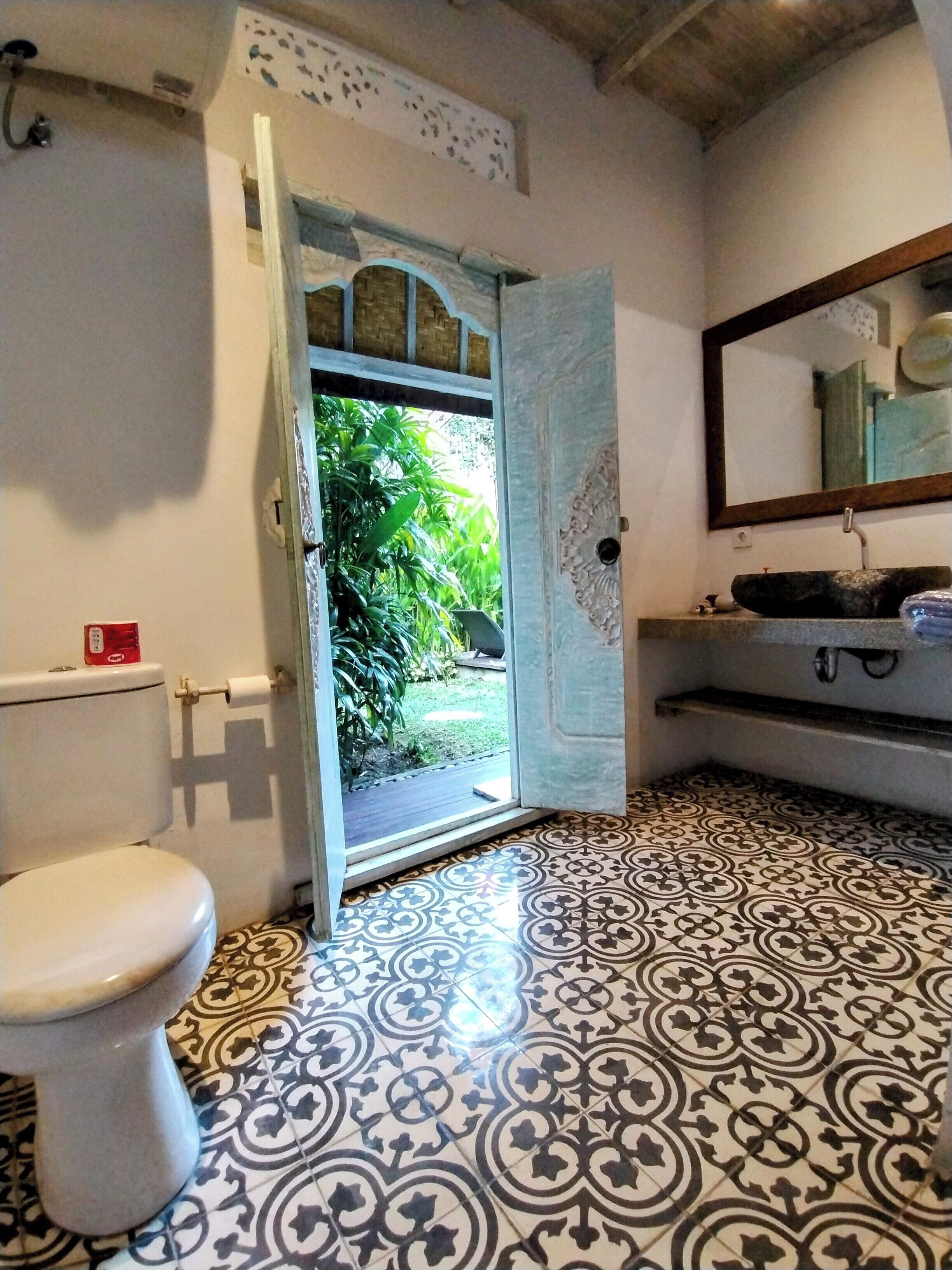 A bathroom with a toilet, a sink with a mirror, patterned floor tiles, and a door leading to an outdoor area with greenery.
