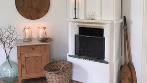 Habitació acollidora amb terra de fusta, una llar de foc blanca, un armari de fusta, un objecte rodó de fusta a la paret i una cistella a terra.