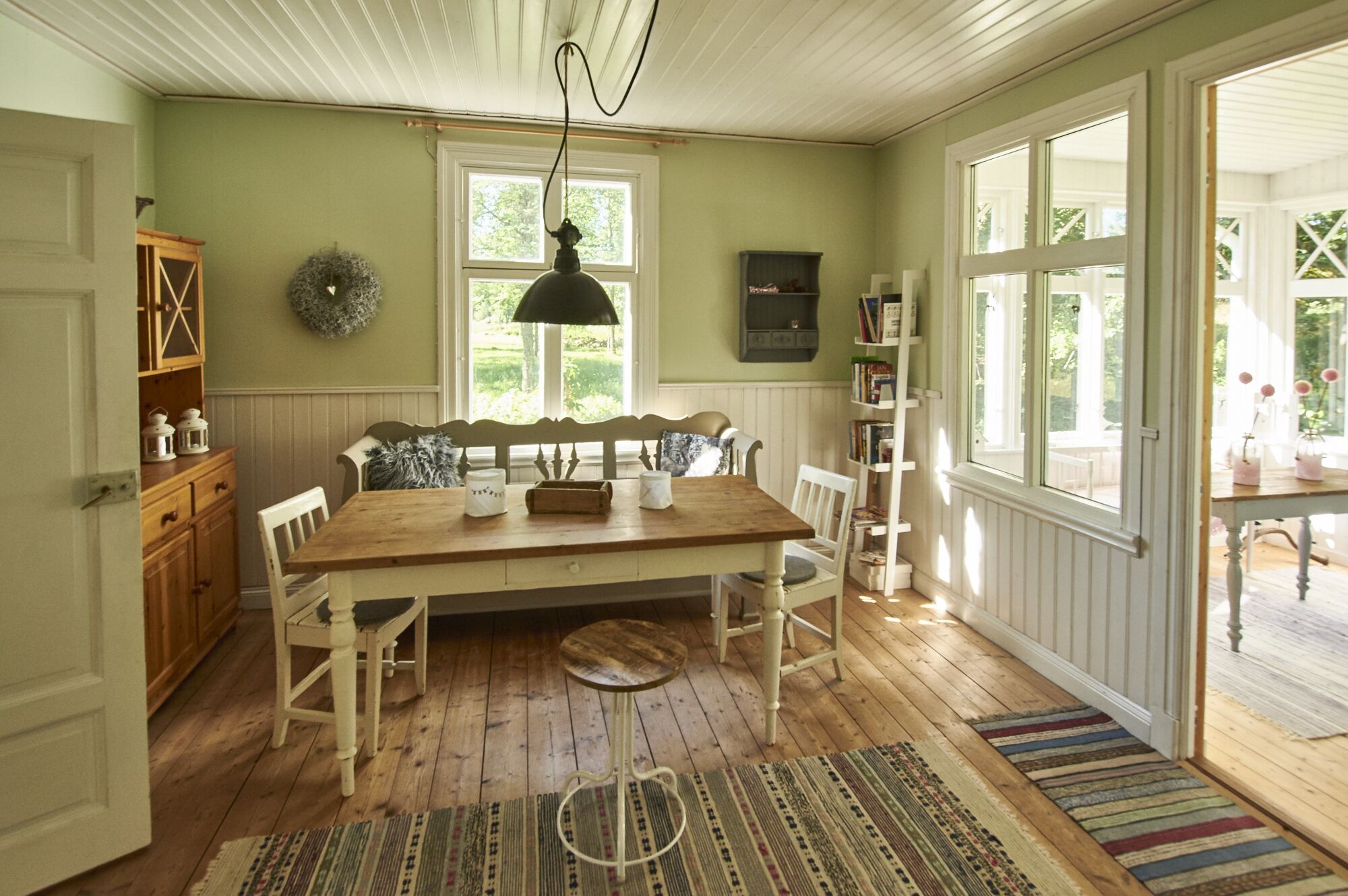 Une salle à manger avec une table en bois, des chaises blanches et un tapis. Une fenêtre offre une vue sur la verdure à l’extérieur.